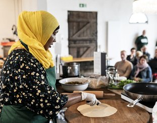 Mørk kvinde i gult tørklæde står foran publikum og laver mad