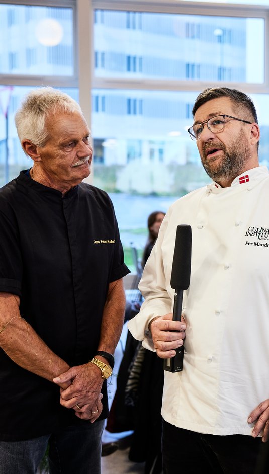 Per Mandrup i hvid kokkejakke med briller står med mikrofon ved siden af dommer, Jens Peter Kolbeck i sort kokkejakke til Kokkenes Kokketalent 2021