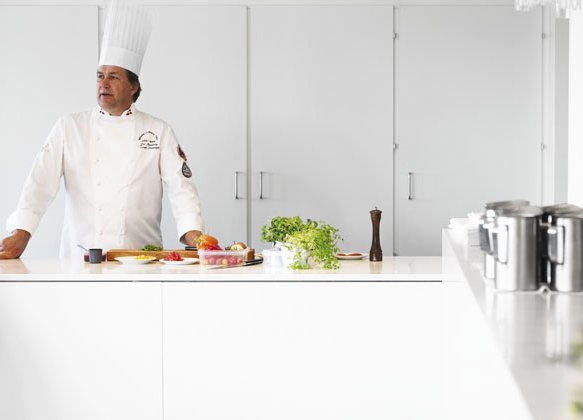 Per Mandrup står i hvidt køkken med hvid kokkejakke og kokkehue med sølv kander, urter, sort peberkværn, skærebræt og forskellige grøntsager